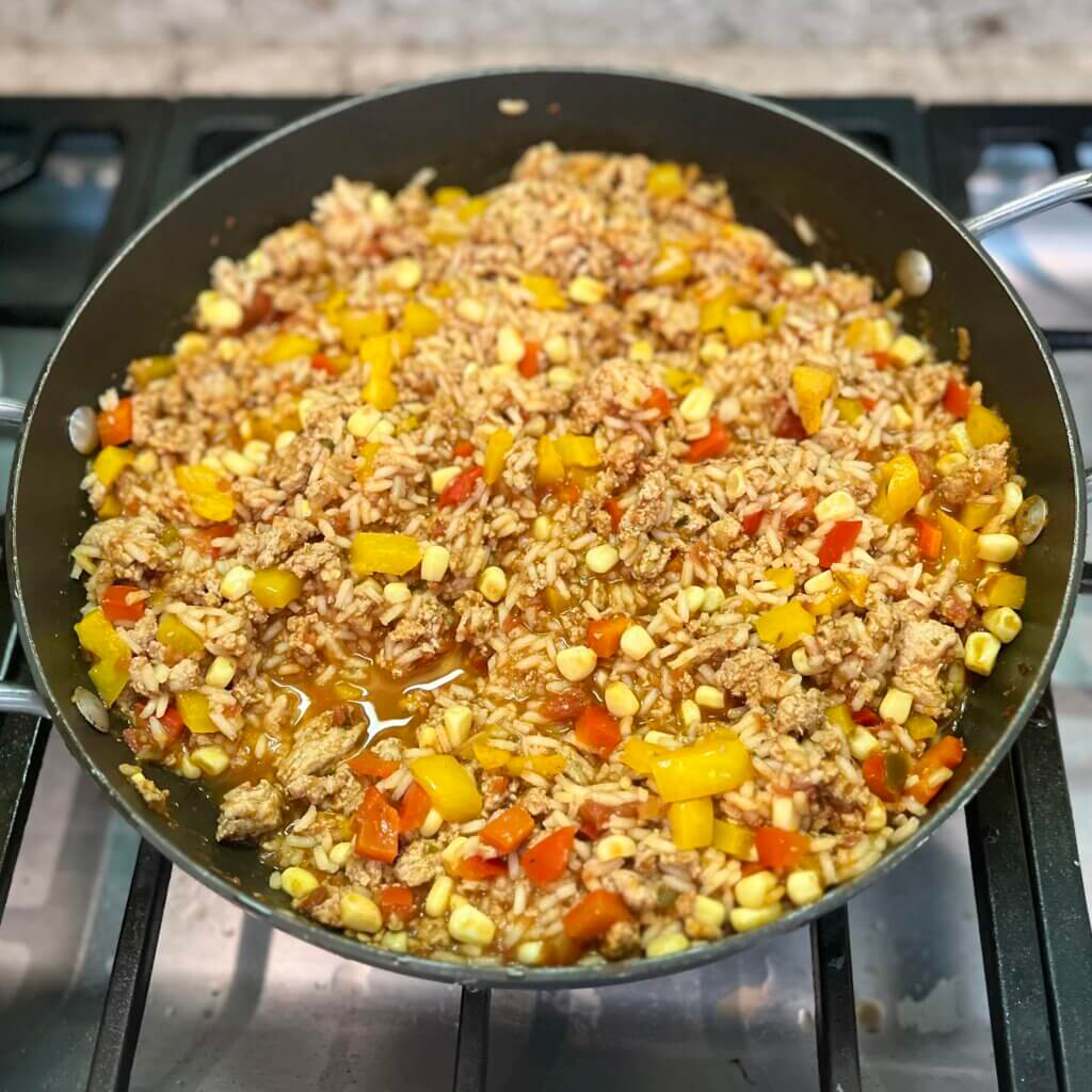 A large frying pan on a stove, filled with minced turkey, rice, chopped bell peppers and corn