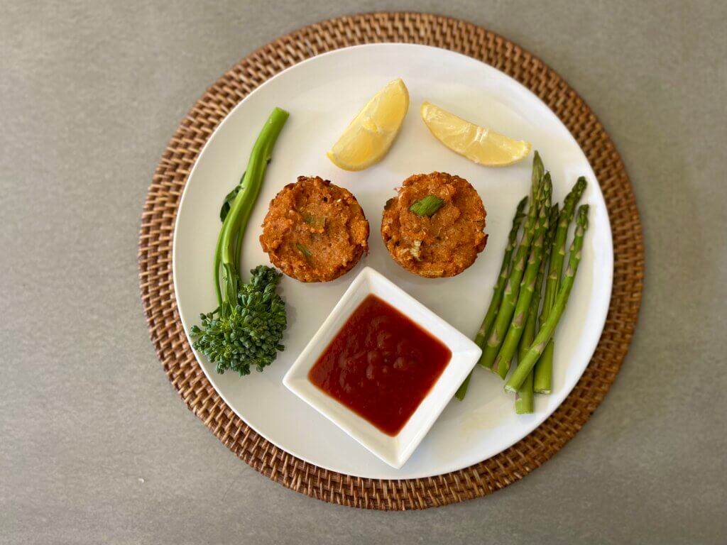 A white plate containing two crab cakes, broccolini, asparagus, lemon wedges and New Canaan Farms Gulf Coast Shrimp Sauce