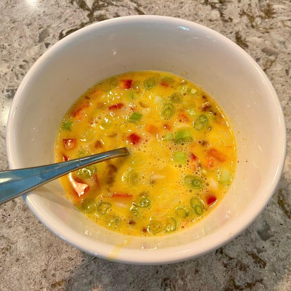 A white bowl with a mixture of whisked raw eggs, red pepper and scallions