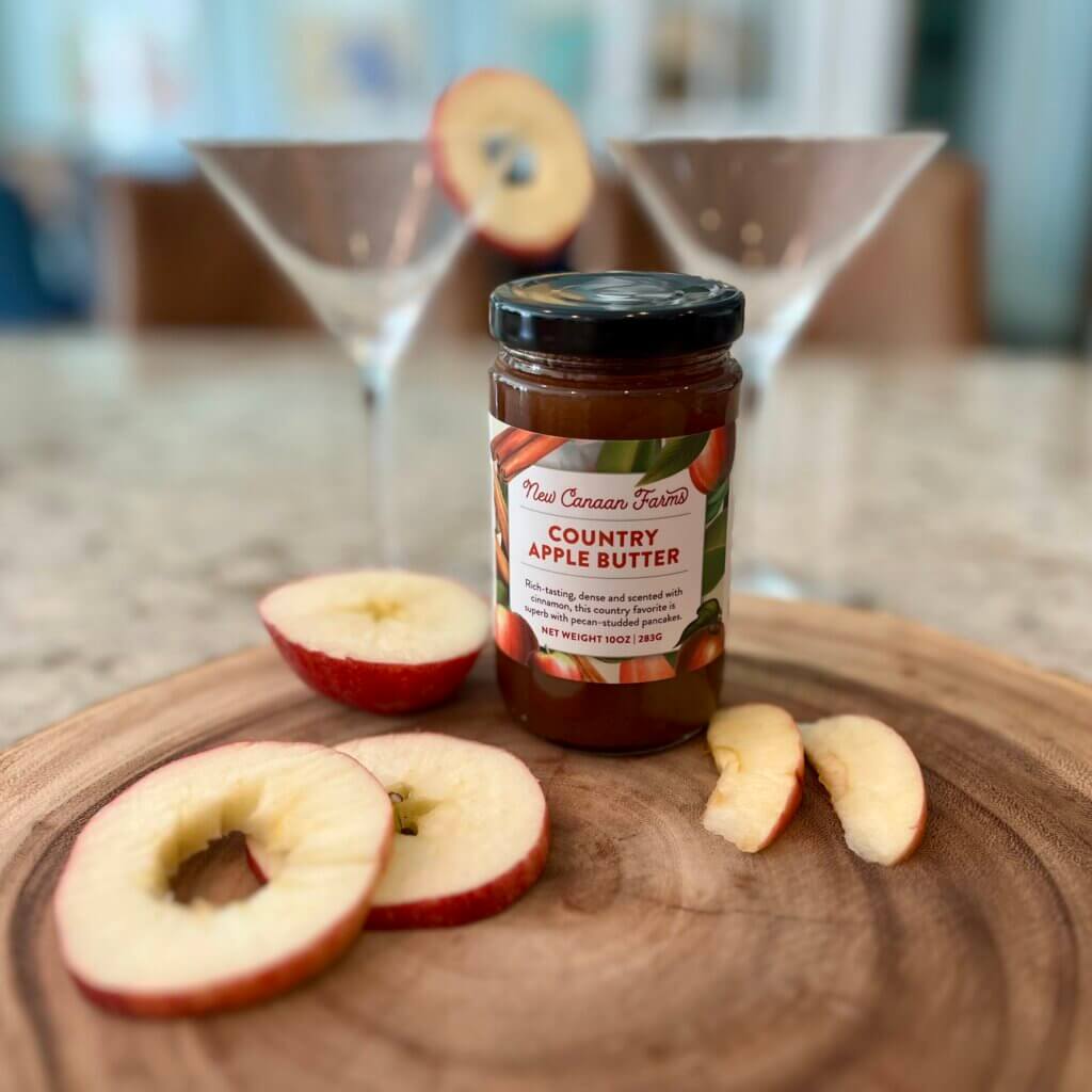 A jar of New Canaan Farms Country Apple butter with a sliced apple and two martini glasses