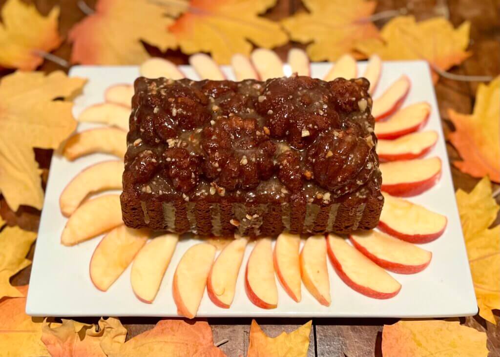 A pumpkin cake with pecan honey glaze, made with New Canaan Farms jams, on a white platter surrounded by apple slices and fall leaves