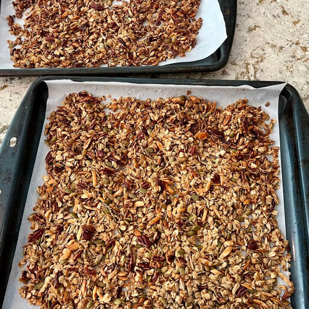Two large baking trays filled with parchment paper and a cooked granola mixture