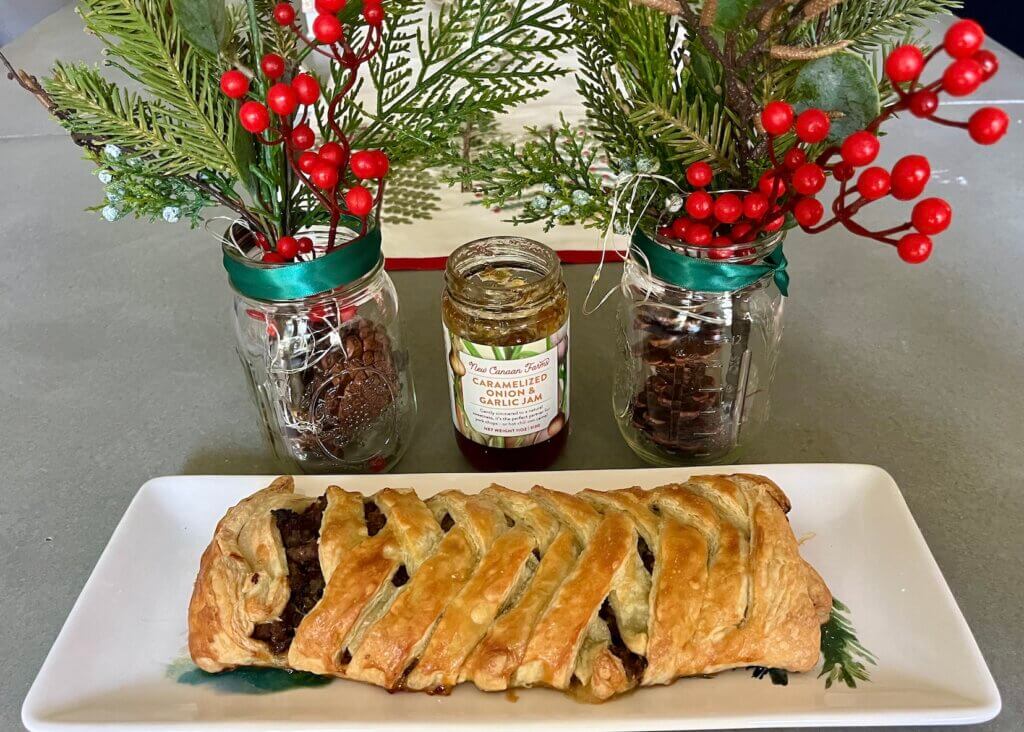 A large festive sausage roll, made with latticed puff pastry, in front of Christmas decorations and a jar of New Canaan Farms Caramelized Onion & Garlic Jam