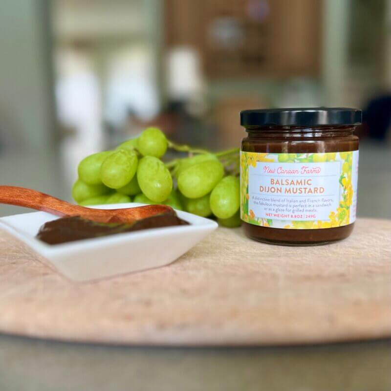 A jar of New Canaan Farms Balsamic Mustard with a small white dish of the mustard and a few green grapes