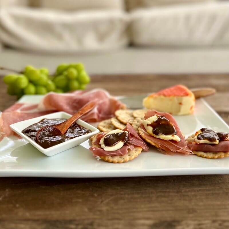 A white platter containing prosciutto, crackers, Chaumes cheese and New Canaan Farms Balsamic Dijon Mustard