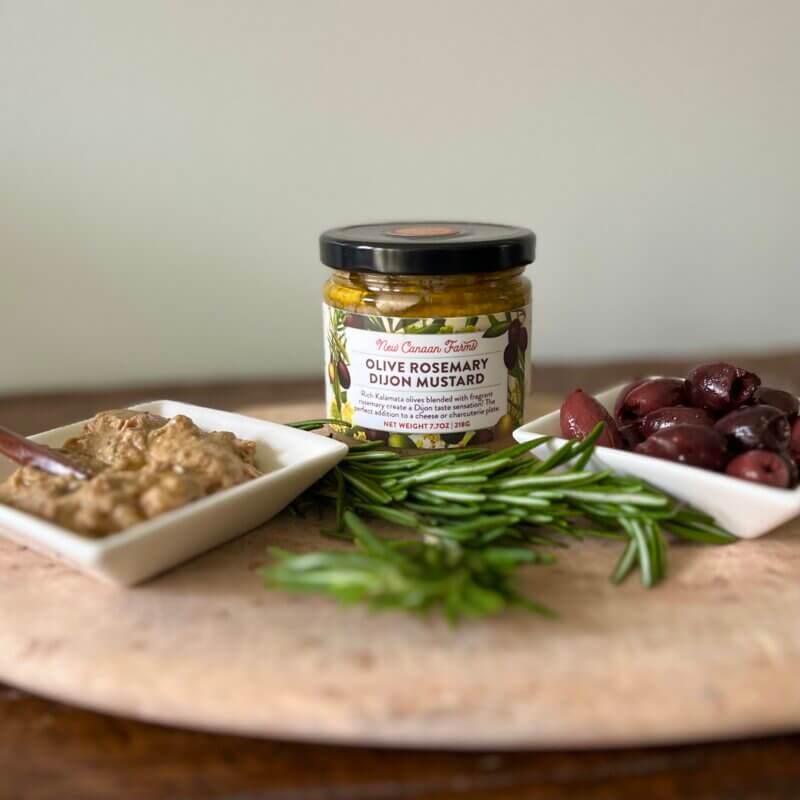 A jar of New Canaan Farms Olive Rosemary Mustard, with a small white dish of mustard, sprigs of rosemary and fresh olives