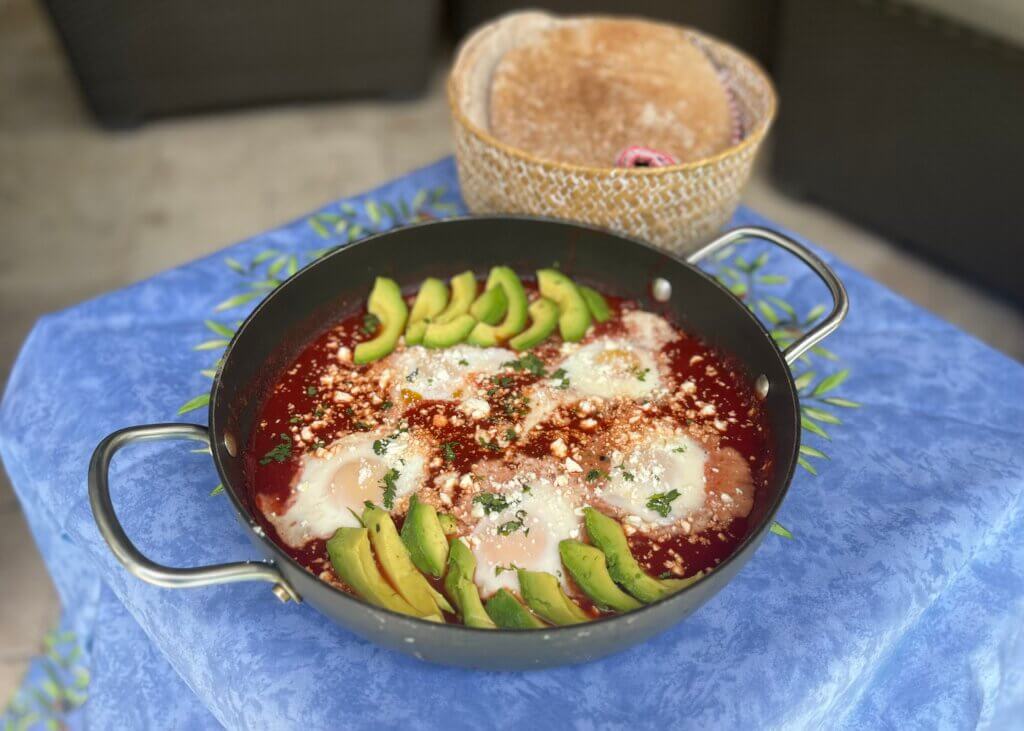 A skillet with 5 baked eggs in New Canaan Farms Pedernales Picante Sauce, garnished with avocado slices and pita bread