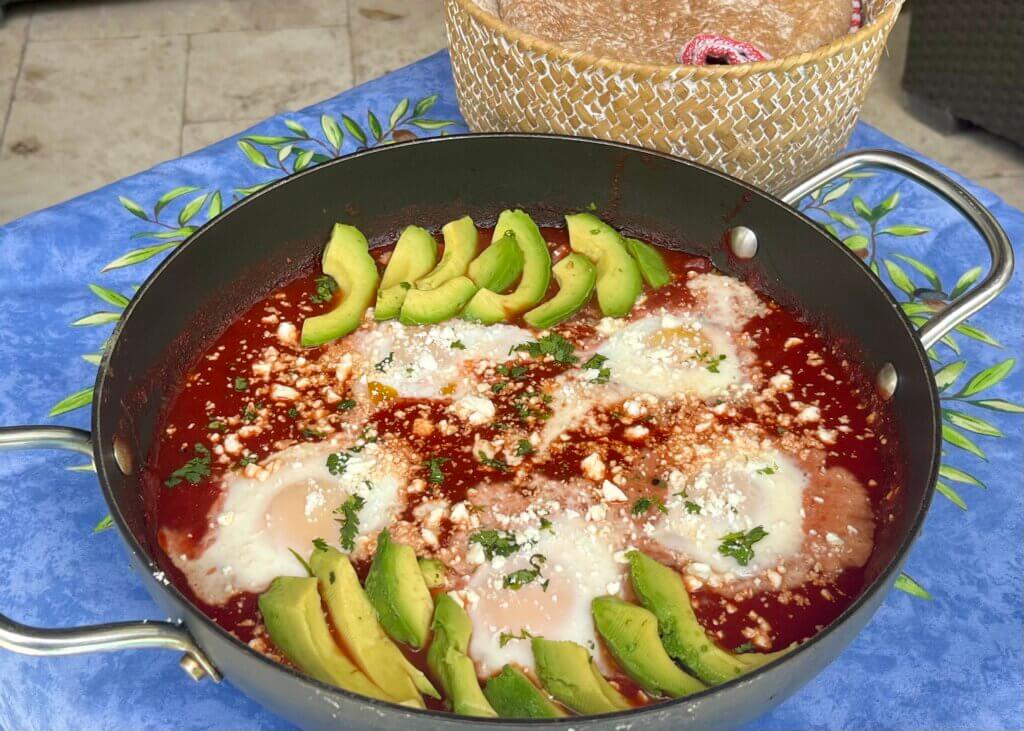 A skillet with 5 baked eggs in New Canaan Farms Pedernales Picante Sauce, garnished with avocado slices and pita bread