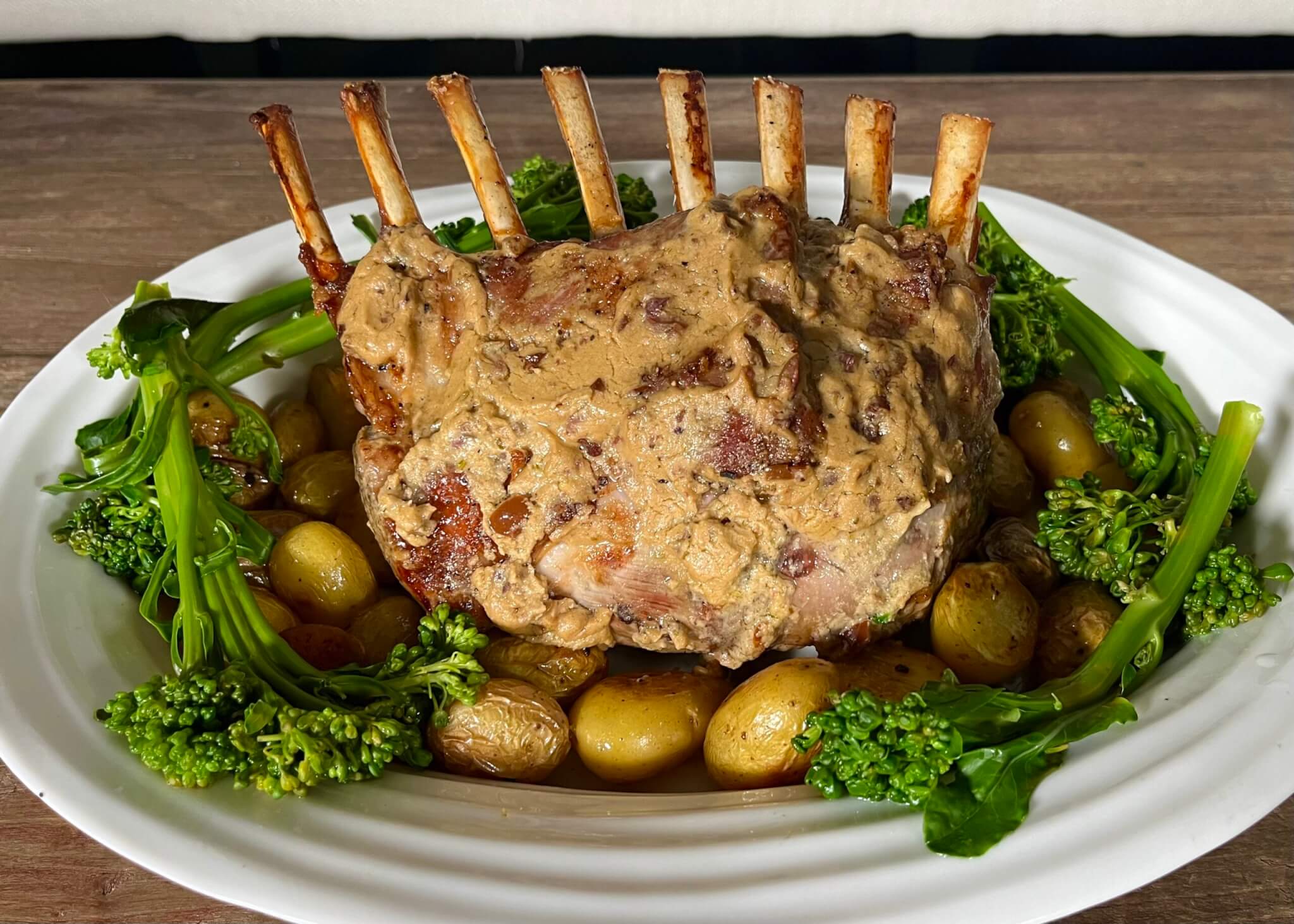 A white platter containing a rack of lamb basted with New Canaan Farms Olive Rosemary Dijon, surrounded by mini potatoes and broccolini